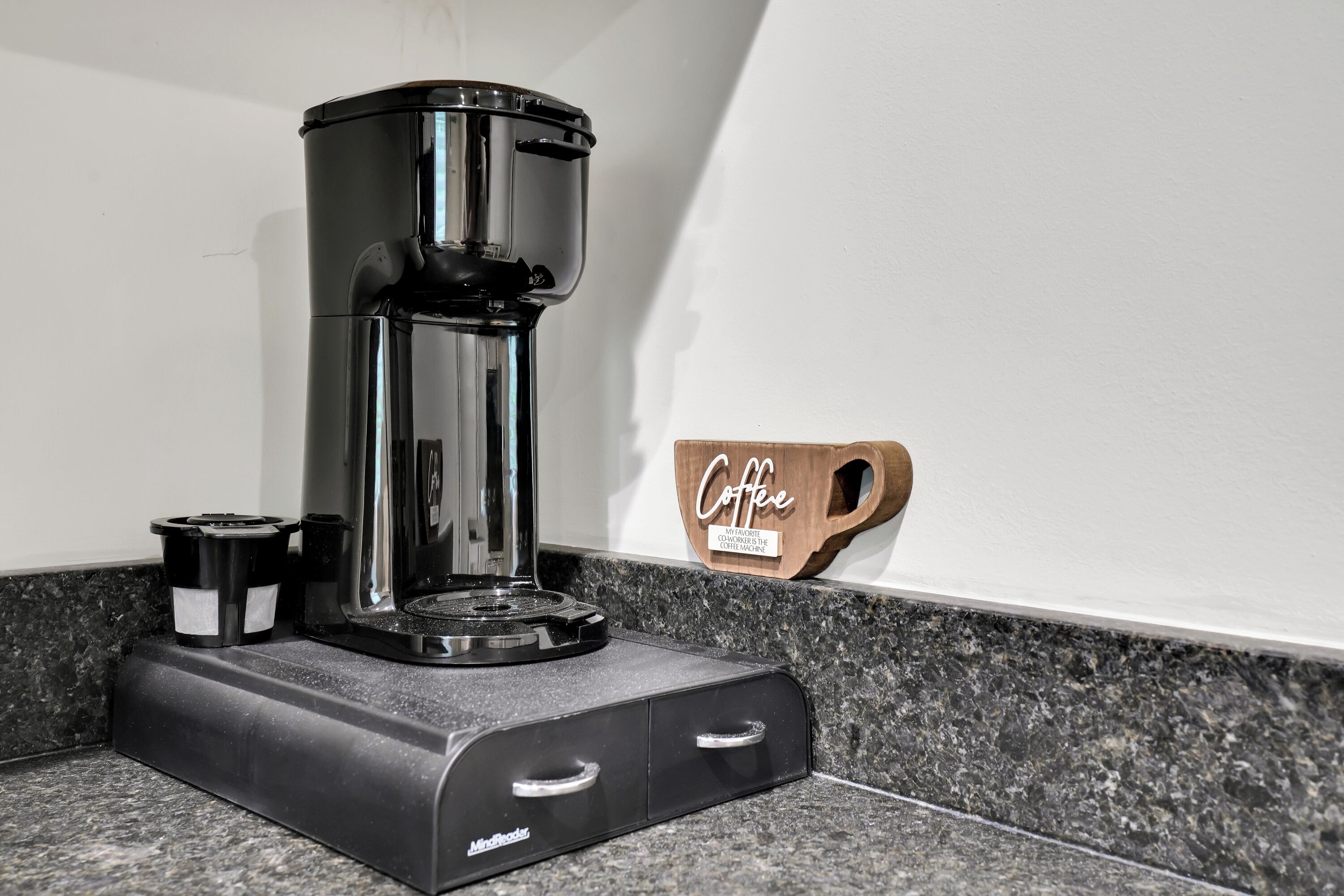 A coffee maker on a countertop with a coffee mug and a wooden sign that says "Coffee".