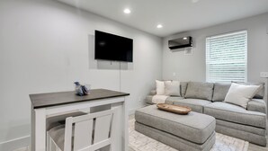 A modern living room with a grey sofa, a white TV stand, and a large window with blinds. - The Jewel – Modern Retreat Near YMCA & Downtown (Wilmington)