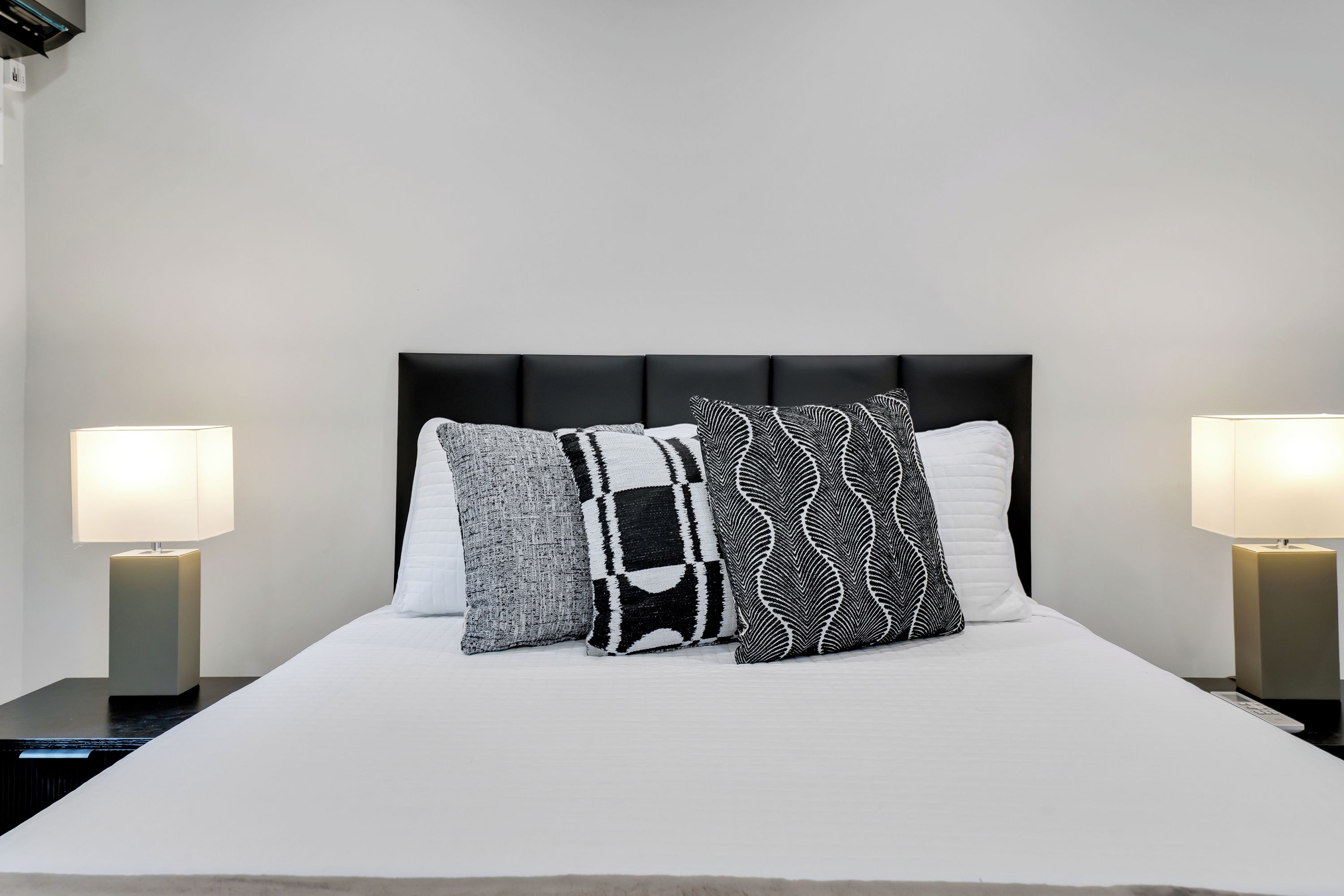 Una cama bien hecha con un cabecero negro, tres almohadas estampadas y dos lámparas de noche a cada lado.