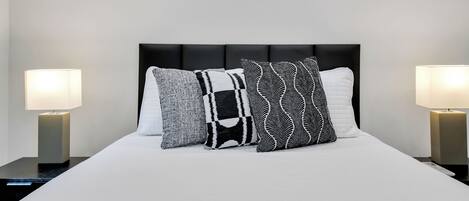 Una cama bien hecha con un cabecero negro, tres almohadas estampadas y dos lámparas de noche a cada lado.