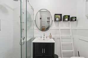 A bathroom with a glass shower enclosure, a white sink with a black cabinet, a round mirror, and three small shelves with potted plants.