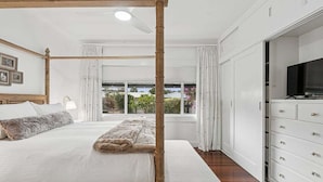 Een slaapkamer met een hemelbed, een groot raam met gordijnen, en een witte dressoir met lades.