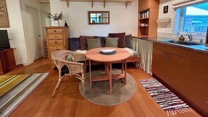A cozy living area with a round table, wicker chair, and a sofa with cushions. There is a ceiling fan, wooden shelves with decorative items, and a window with a view.