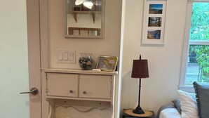 A bedroom with a bed, a bedside table with a lamp, a small wooden stool with towels, a dresser with a mirror, and a window with a view of greenery.