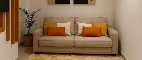 A living room with a beige sofa, two orange cushions, a fluffy beige rug, a potted plant, and two wall-mounted artworks.
