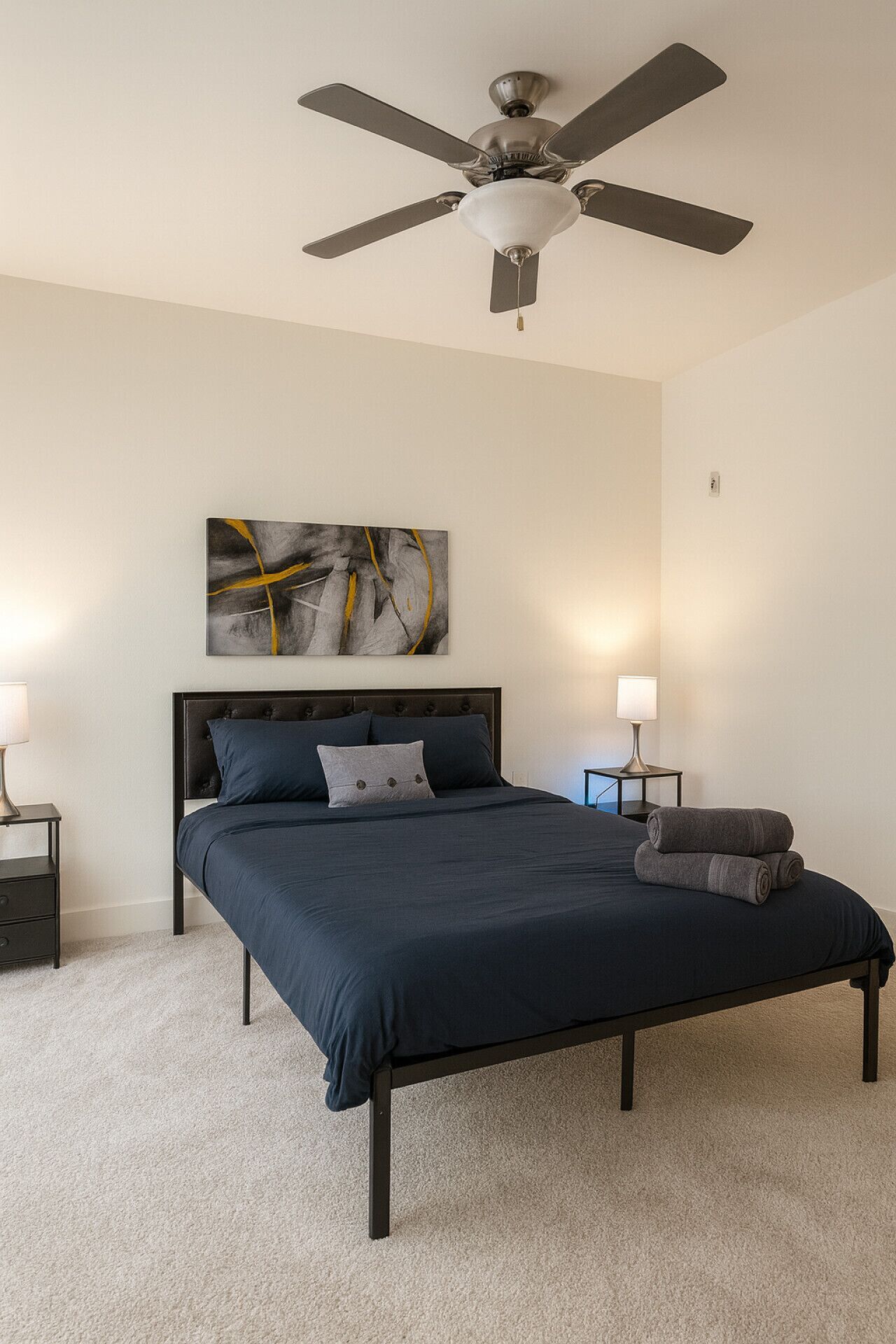 A bedroom with a bed, two bedside tables, a ceiling fan, and a wall-mounted abstract painting.