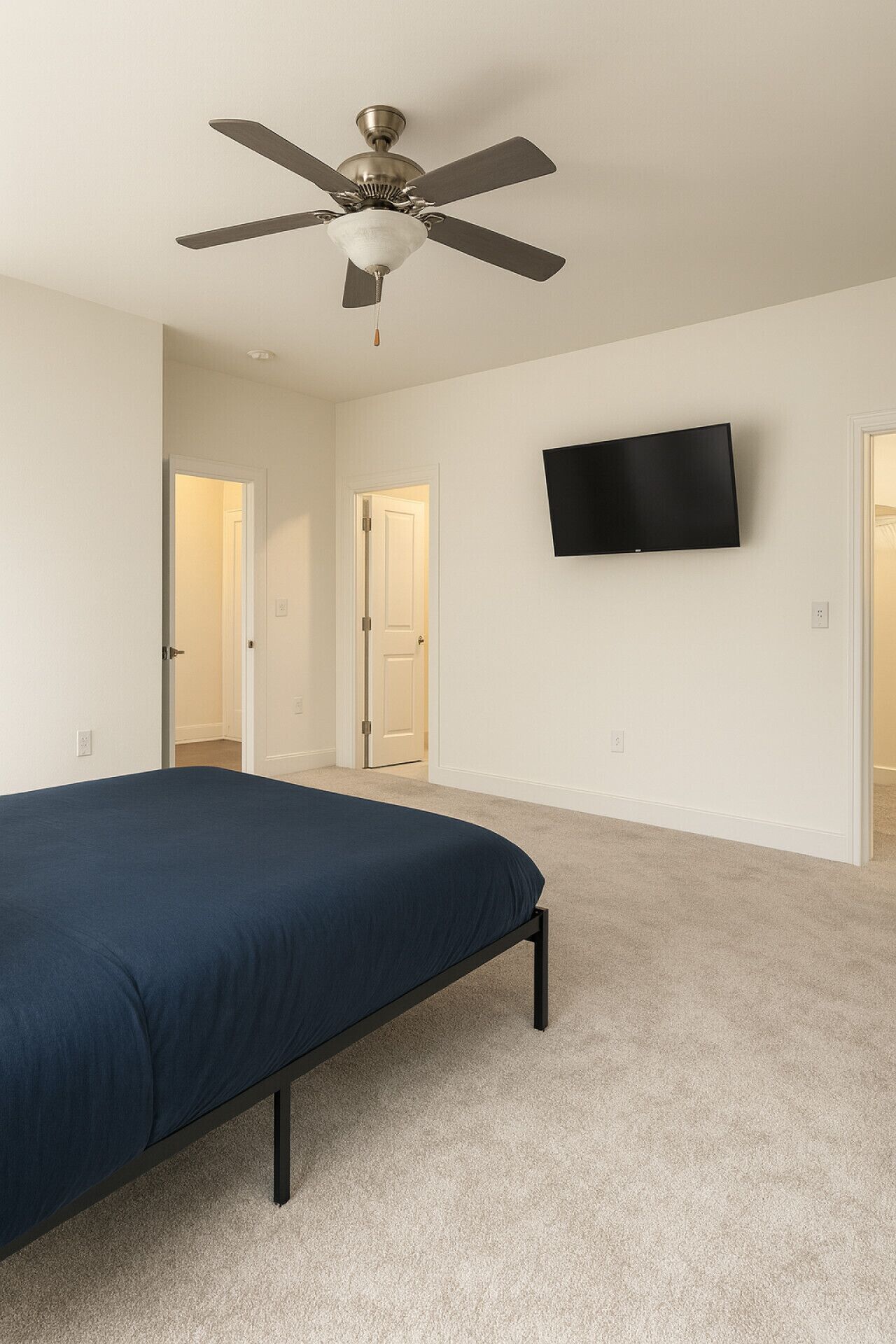 Une chambre à coucher comprenant un lit, un ventilateur de plafond, un téléviseur fixé au mur et deux portes donnant sur d’autres pièces.