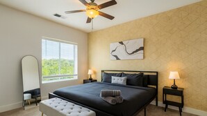 A bedroom with a large bed, two bedside tables with lamps, a bench, a ceiling fan, and a window with blinds.