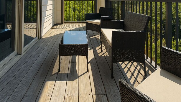 A covered outdoor deck with wooden flooring, a glass door entrance, and a view of a green landscape.