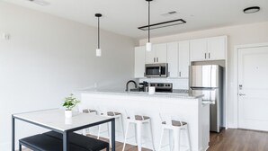 A modern kitchen with white cabinets, a stainless steel refrigerator, a microwave, and a built-in oven. There is a dining table with four white chairs and a black bench.