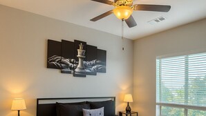 Une chambre avec un grand lit, deux tables de chevet avec des lampes, un ventilateur de plafond et une fenêtre avec des stores.