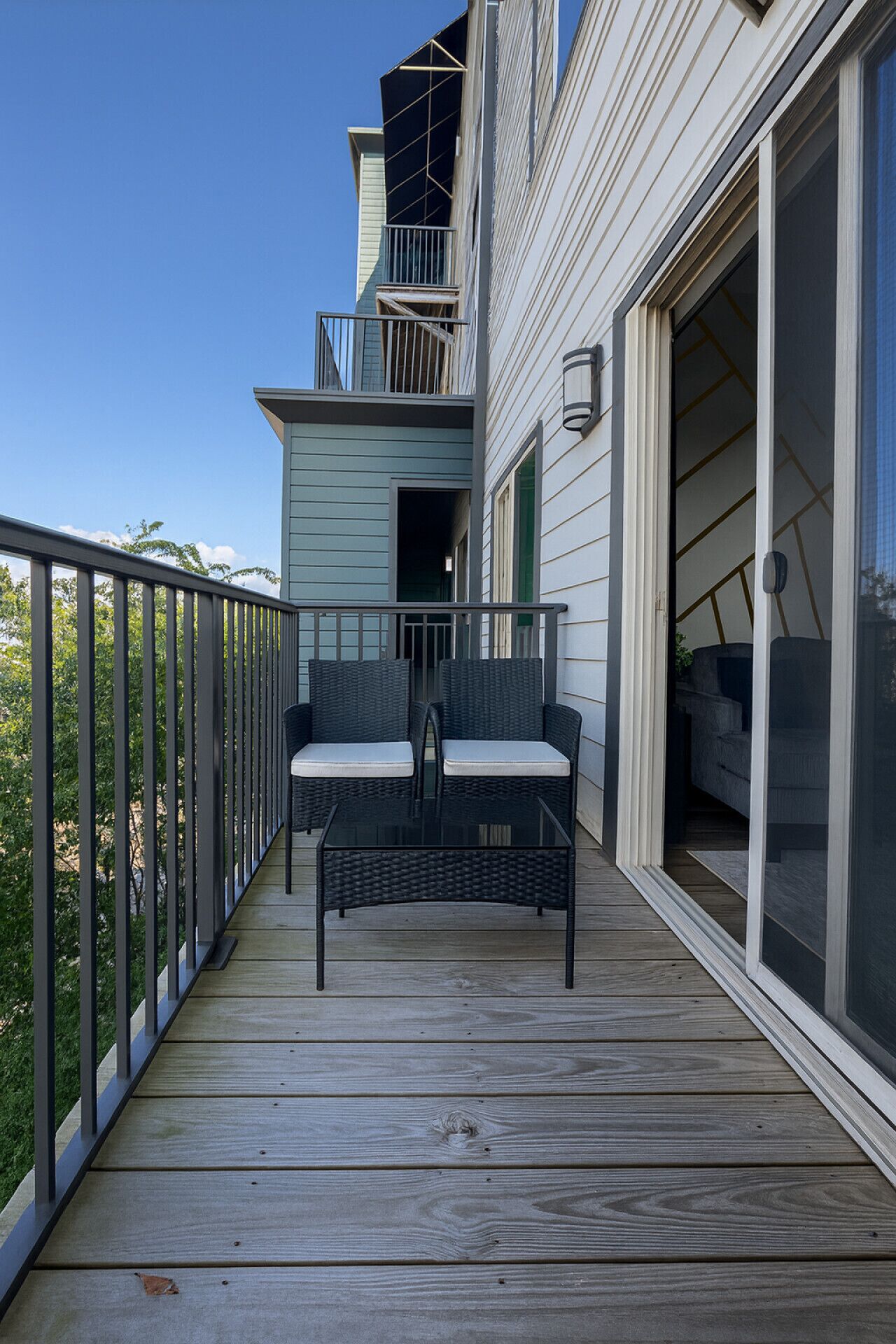 A wooden deck with a black wicker sofa and cushions, a glass door leading to a covered area, and a metal railing.