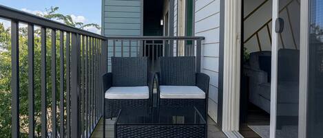 A wooden deck with a black wicker sofa and cushions, a glass door leading to a covered area, and a metal railing.