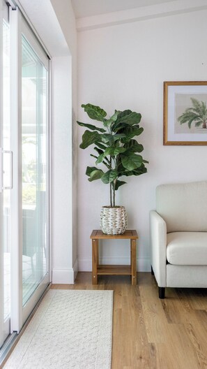 Un salon avec une grande plante posée sur un support en bois, un tableau encadré accroché au mur et une porte-fenêtre coulissante.