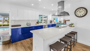 Een moderne keuken met een centraal eiland, blauwe kasten, witte aanrechtbladen en een grote klok aan de muur.