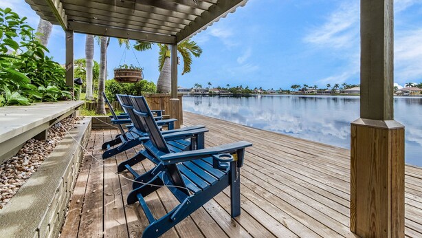 Een houten terras met blauwe stoelen en een hangstoel, met uitzicht op een rustig wateroppervlak, omgeven door groen en palmbomen.