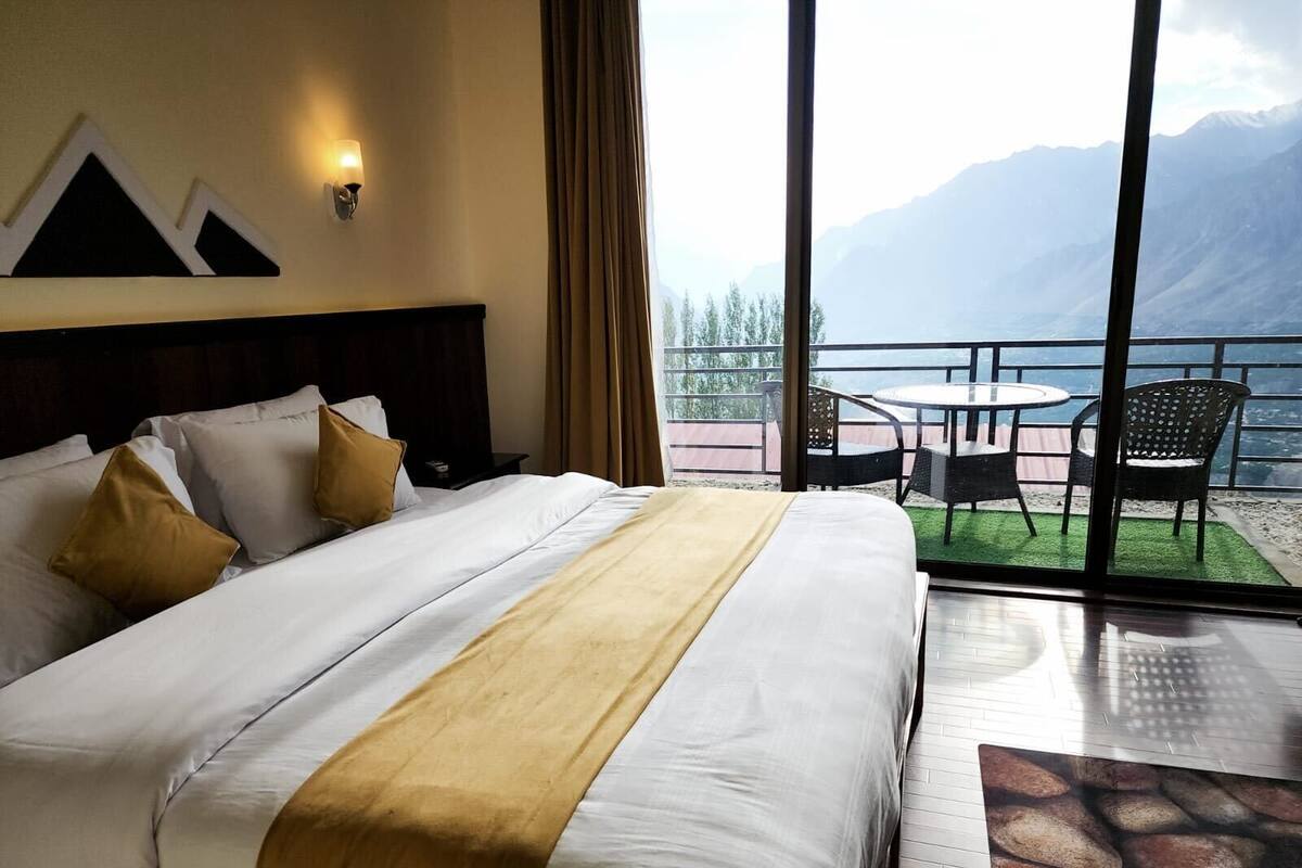 Una habitación de hotel con una cama grande, un balcón con mesa y sillas, y vistas a las montañas.