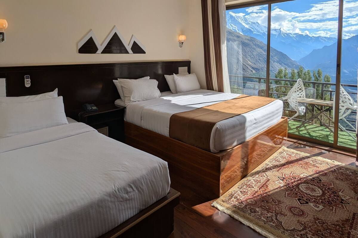 Habitación de hotel con dos camas, ventilador de techo, ventana grande con vista a la montaña y alfombra estampada.