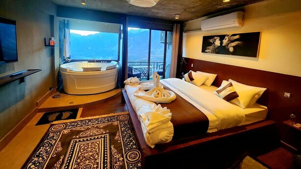 A modern hotel room with a large bed, a jacuzzi, a TV, and a balcony with a mountain view. - HARD ROCK HUNZA (Karimabad)