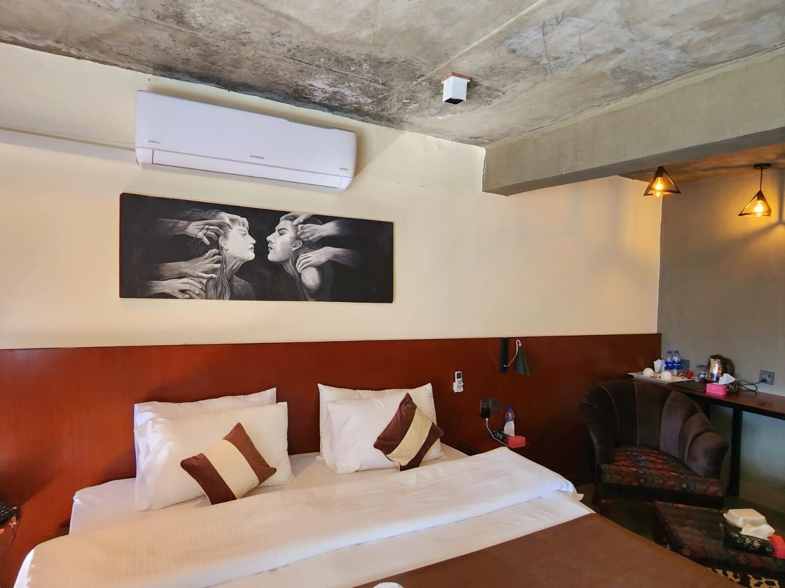 A hotel room with a large bed, a headboard, an air conditioning unit, a wall-mounted artwork, a desk with a chair, and a wall-mounted lamp.