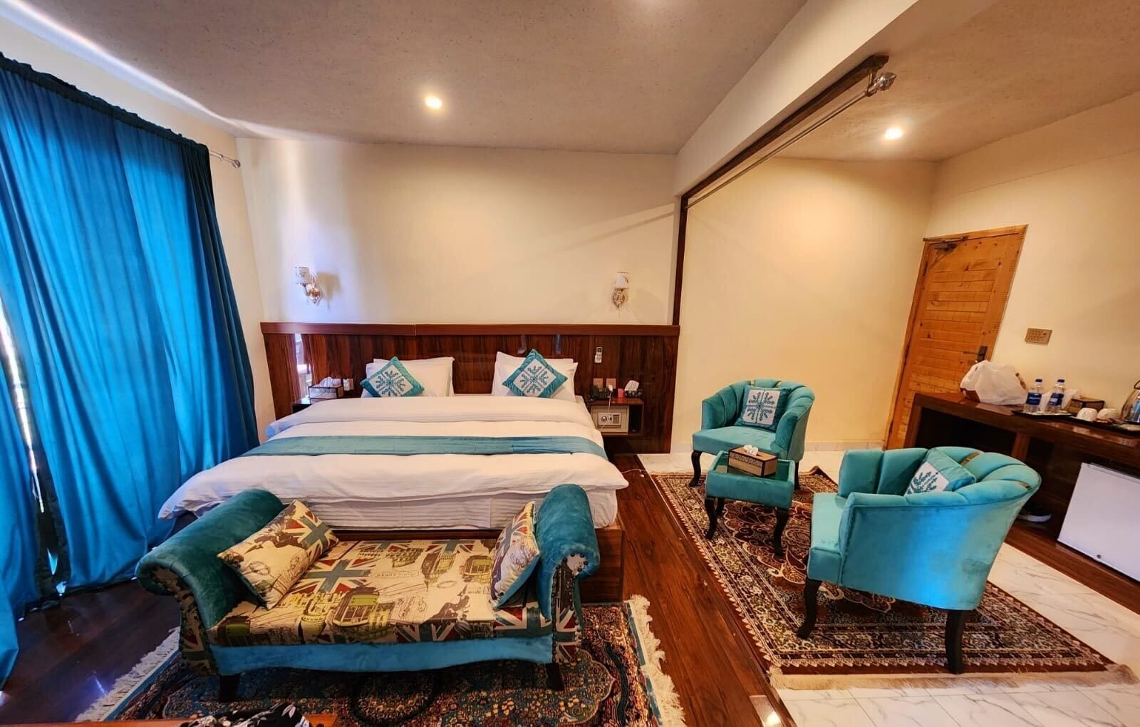 A hotel room with a large bed, two armchairs, a desk with a computer, and a wooden door.