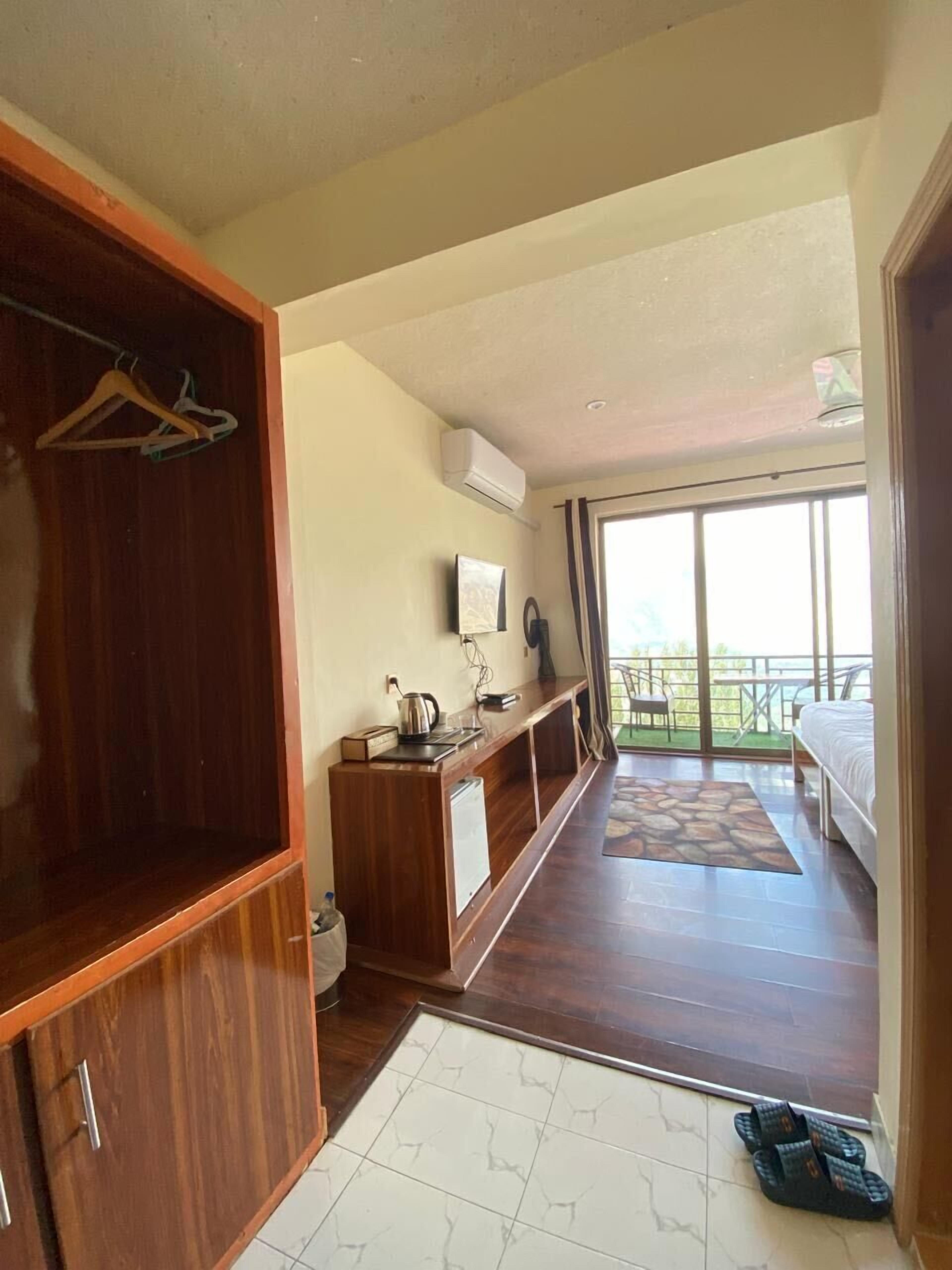 A bedroom with a wooden wardrobe, a small kitchenette, a balcony with a table and chairs, and a bed.