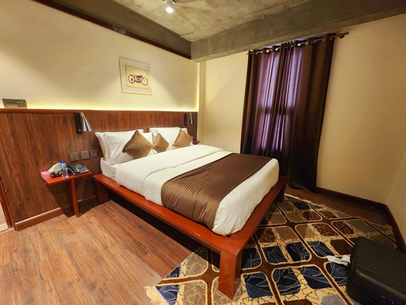 A hotel room with a wooden bed, a nightstand with a lamp, a wall-mounted phone, and a patterned rug.