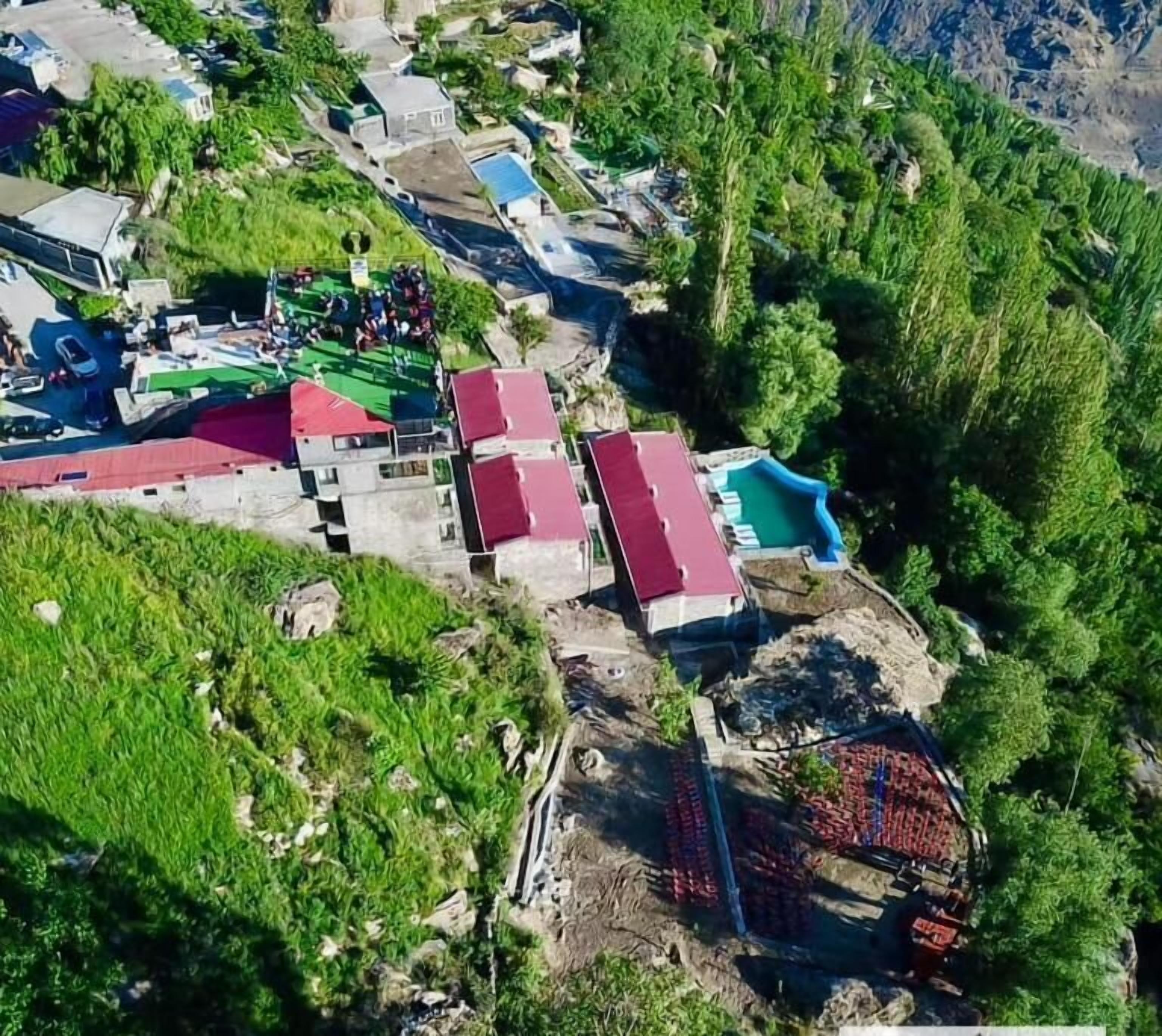 An aerial view of a residential area with multiple buildings, some with red roofs, surrounded by lush greenery and a mountainous backdrop.
