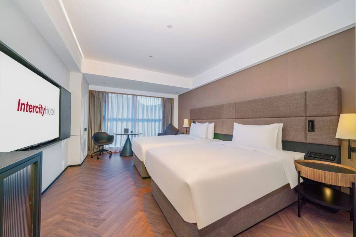 Ein Hotelzimmer mit einem großen Bett, einem Schreibtisch, einem Stuhl und einem Fernseher, der den Namen des Hotels anzeigt.