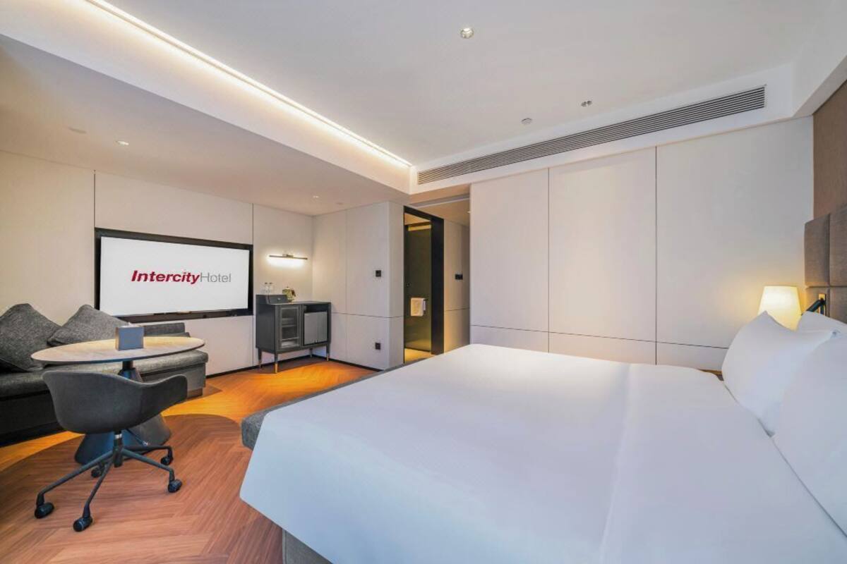 Ein modernes Hotelzimmer mit Bett, Schreibtisch, Sessel, Sofa und einem Fernseher, der 'Intercity Hotel' anzeigt.