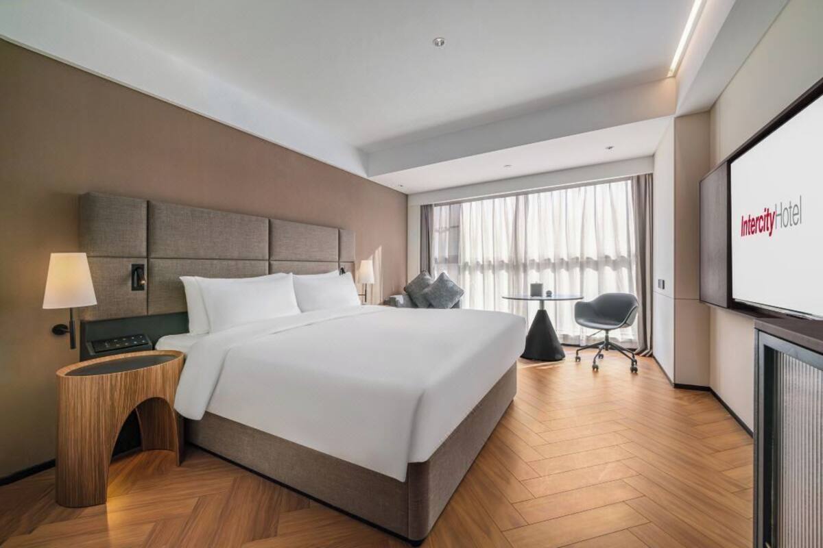 Ein Hotelzimmer mit einem großen Bett, einem Nachttisch, einem Sessel, einem Schreibtisch und einem Fernseher, der den Namen des Hotels anzeigt.
