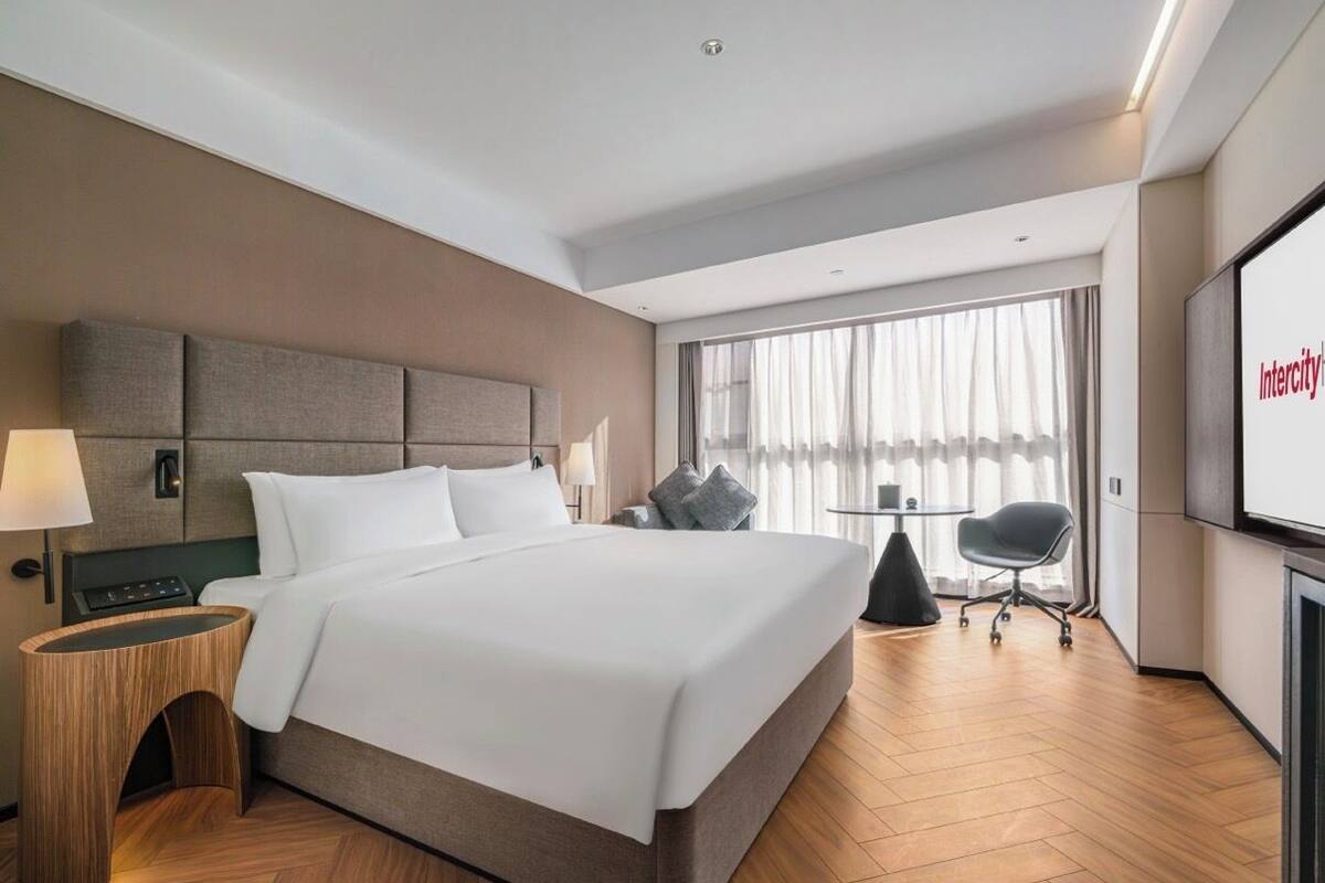 Ein modernes Hotelzimmer mit einem großen Bett, einem Nachttisch, einem Sessel, einem Schreibtisch und einem Fernseher.