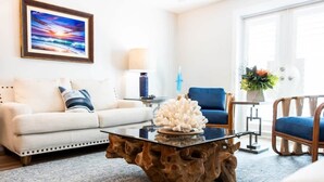 A living room with a white sofa, blue armchairs, a glass coffee table with a coral centerpiece, and a framed sunset painting on the wall.