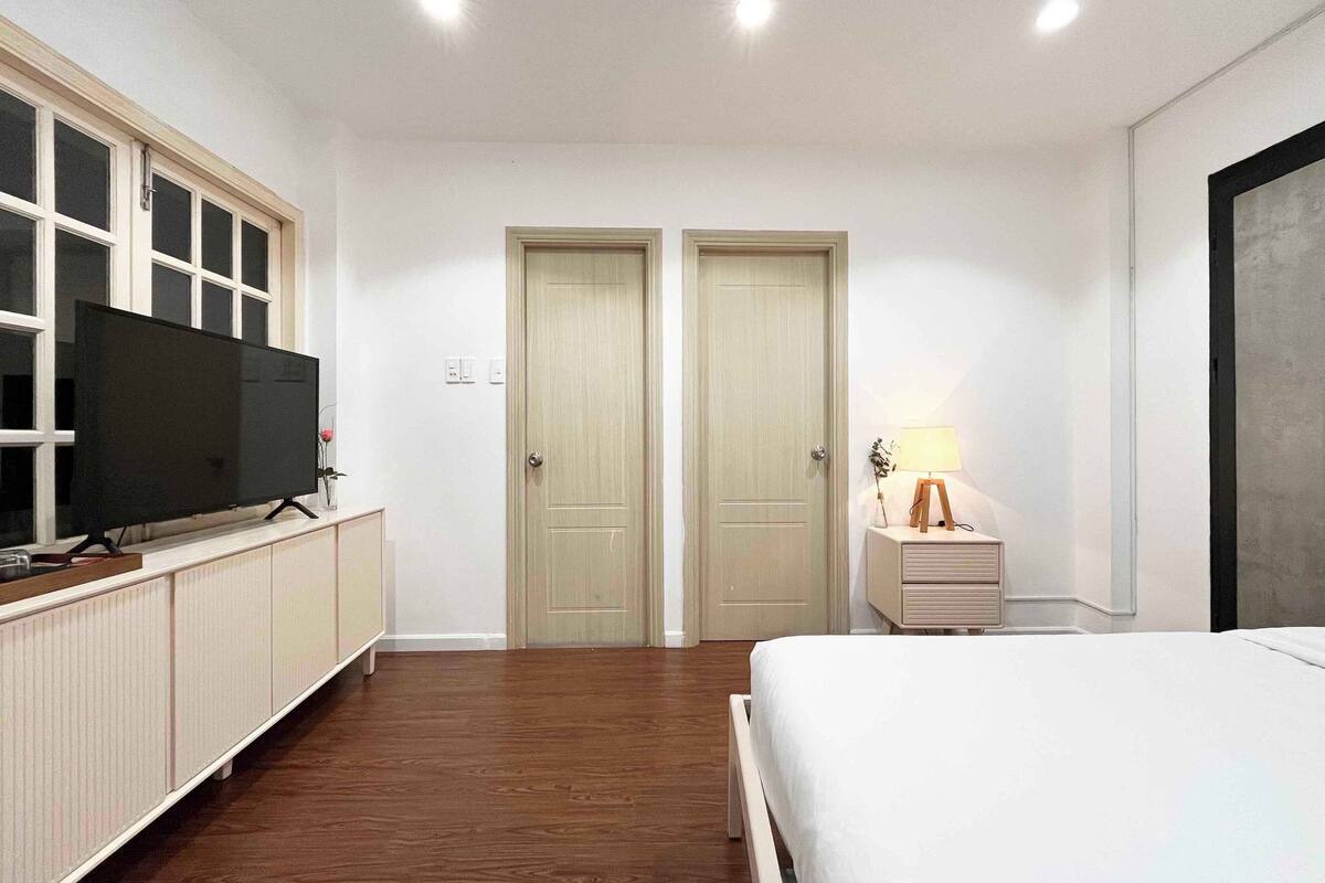 A bedroom with a bed, a TV on a stand, two doors, a bedside table with a lamp, and a radiator.