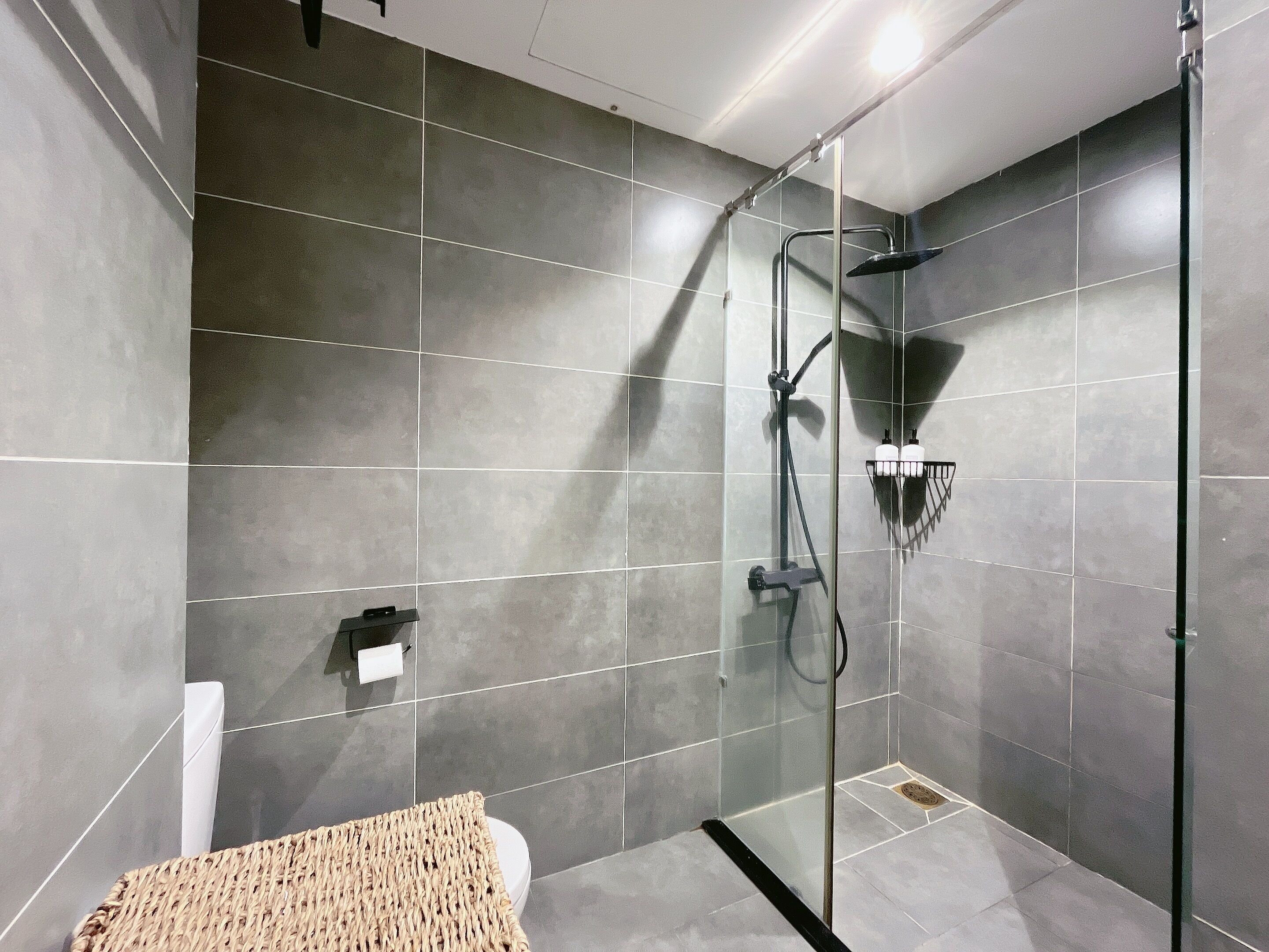 A modern bathroom with a glass-enclosed shower, grey tiles, and a woven bath mat.
