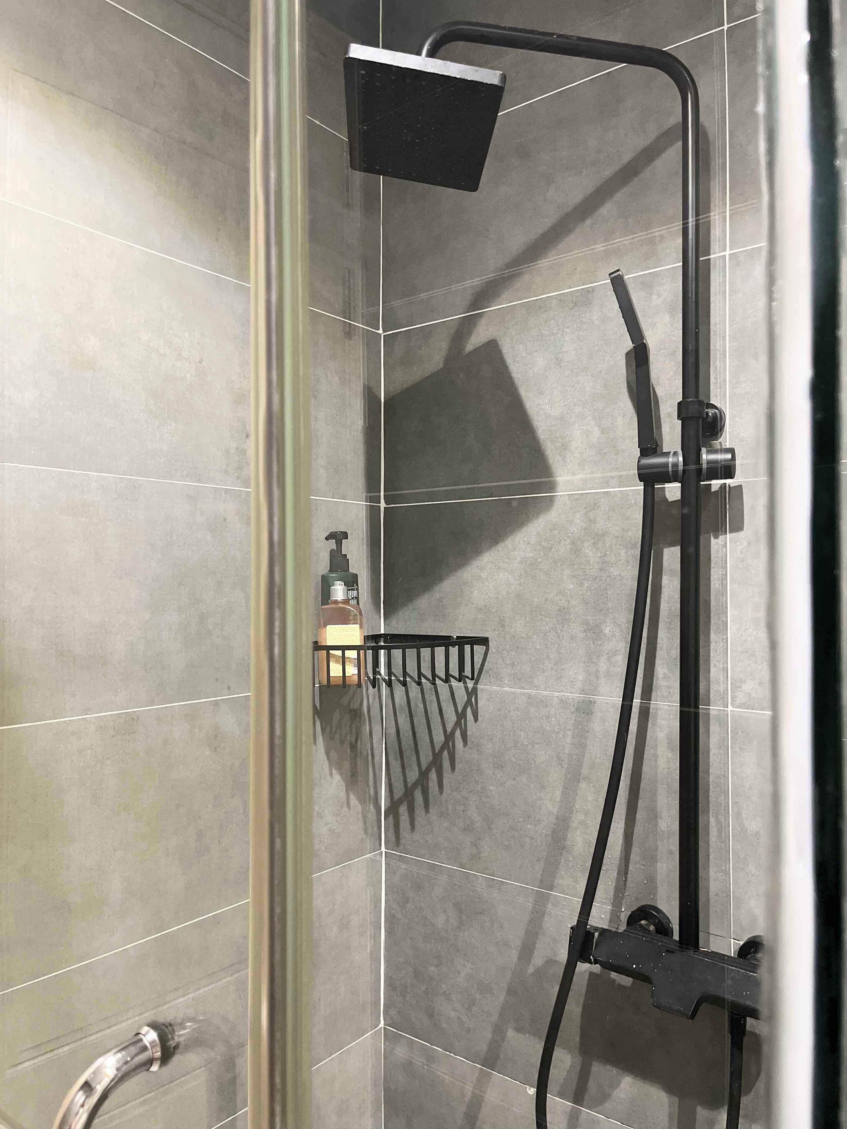 A shower area with a handheld showerhead, a wall-mounted faucet, and a soap dispenser on a shelf.