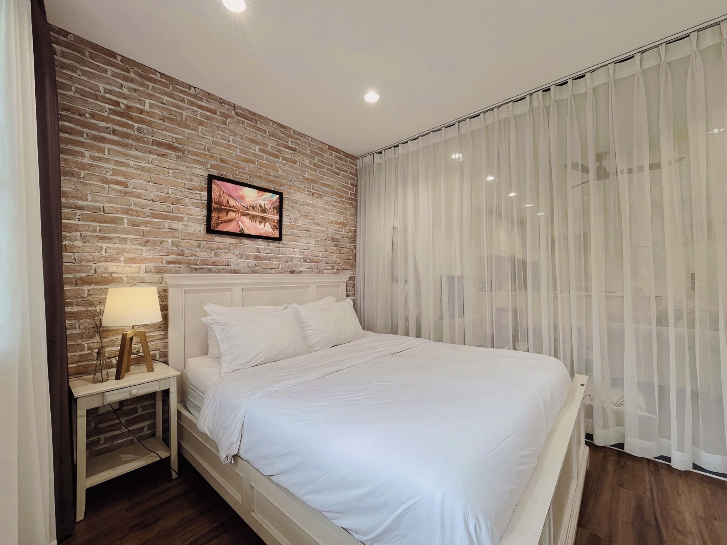 A bedroom with a brick-patterned wall, a bed with white bedding, a nightstand with a lamp, and sheer curtains.
