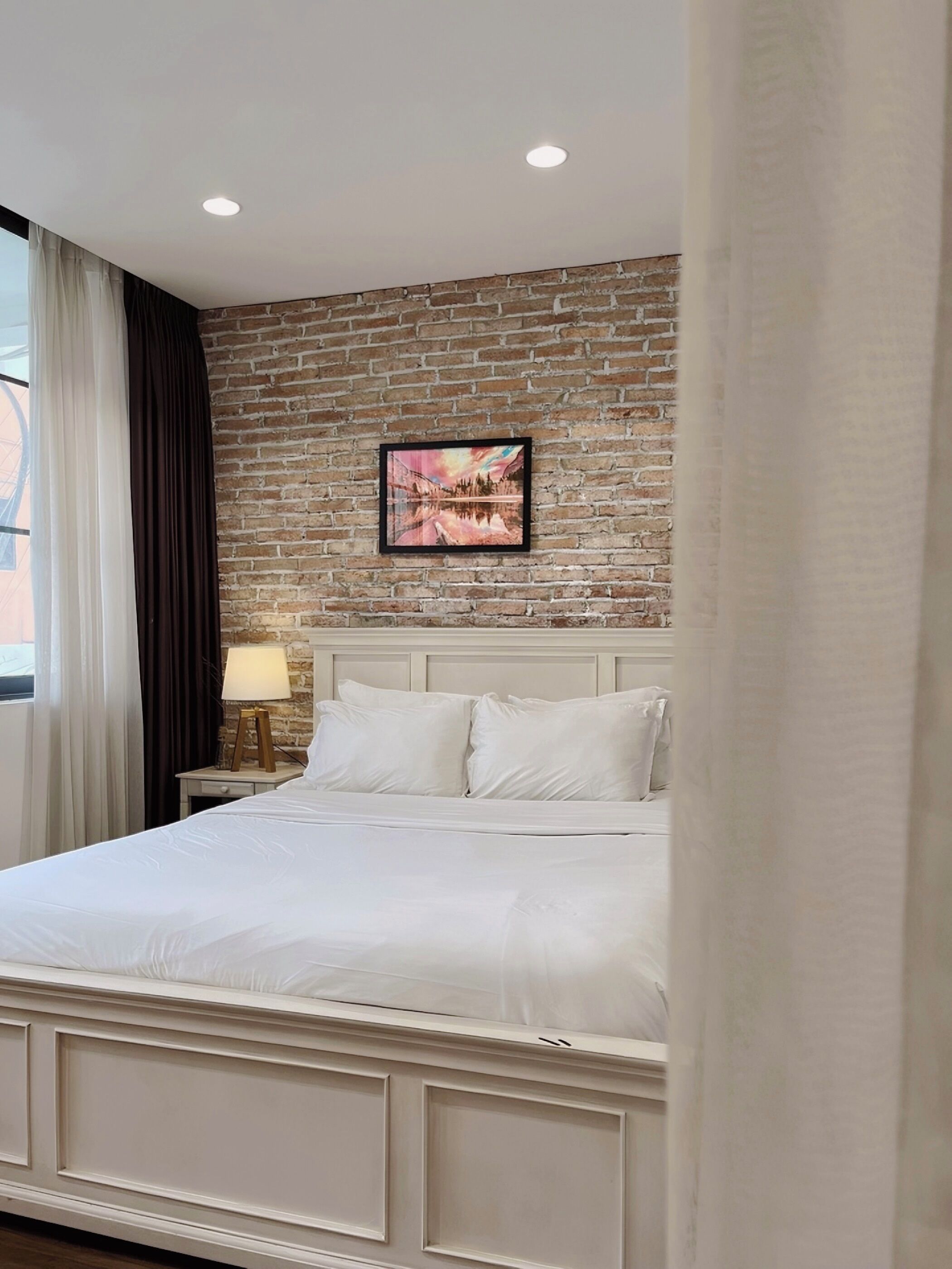 A hotel room with a brick wall, a bed with white bedding, a nightstand with a lamp, and a framed picture on the wall.