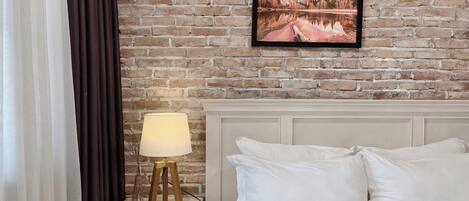 A hotel room with a brick wall, a framed picture of a landscape, a bedside lamp, and a bed with white bedding.