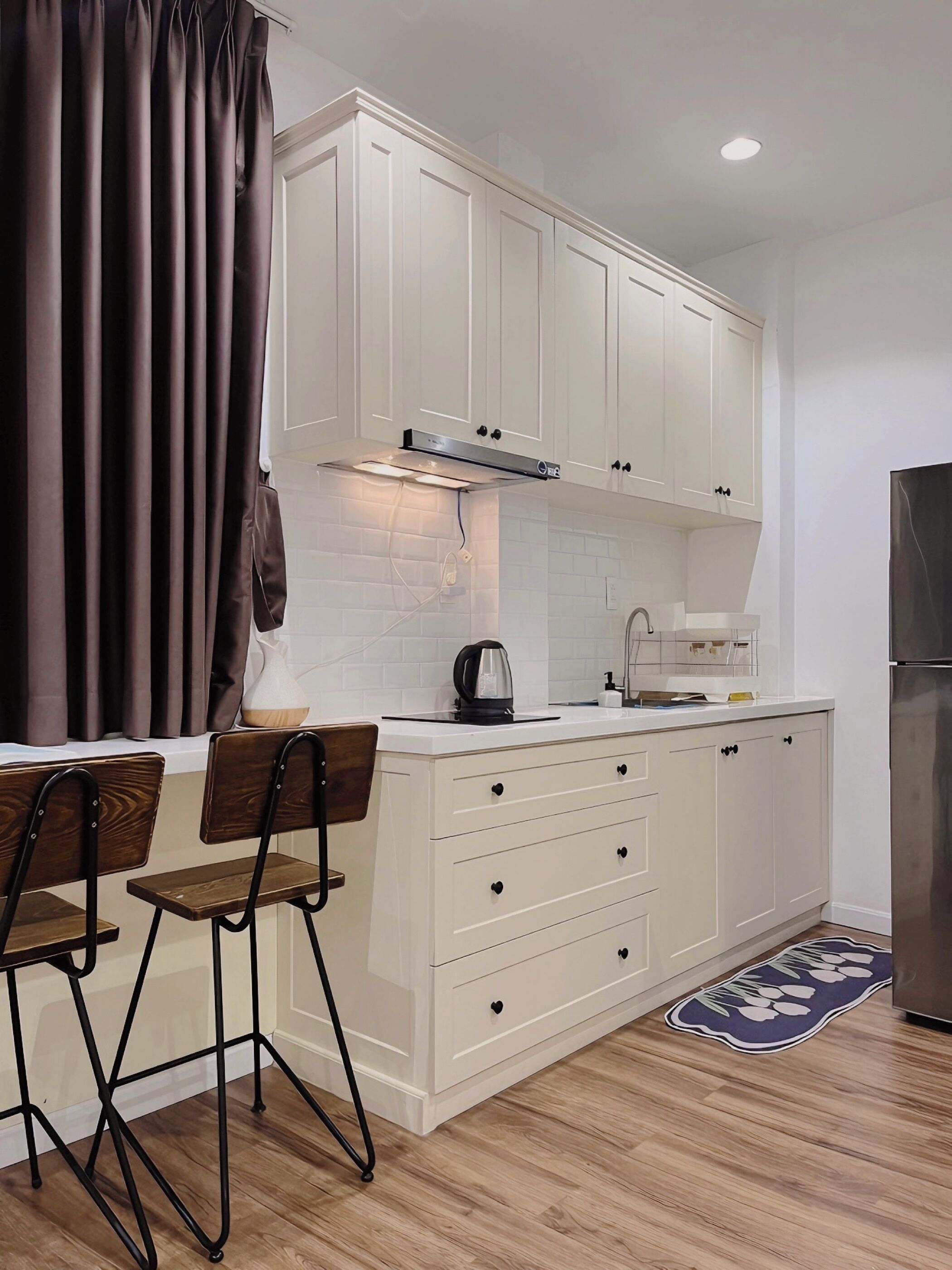 A modern kitchen with white cabinets, a stainless steel range hood, a kettle, and a refrigerator.
