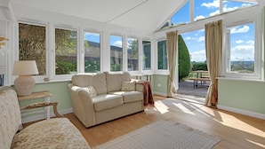 A cozy living room with a sofa, a table, and a lamp. Large windows offer a view of the outdoors. - Whitehall Cottage - sleeps 8 guests  in 4 bedrooms (Painswick)