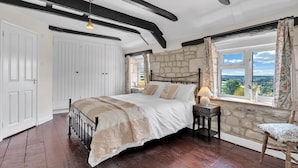 A bedroom with a stone wall, a large bed with white bedding, a nightstand with a lamp, and a chair by the window. - Whitehall Cottage - sleeps 8 guests  in 4 bedrooms (Painswick)