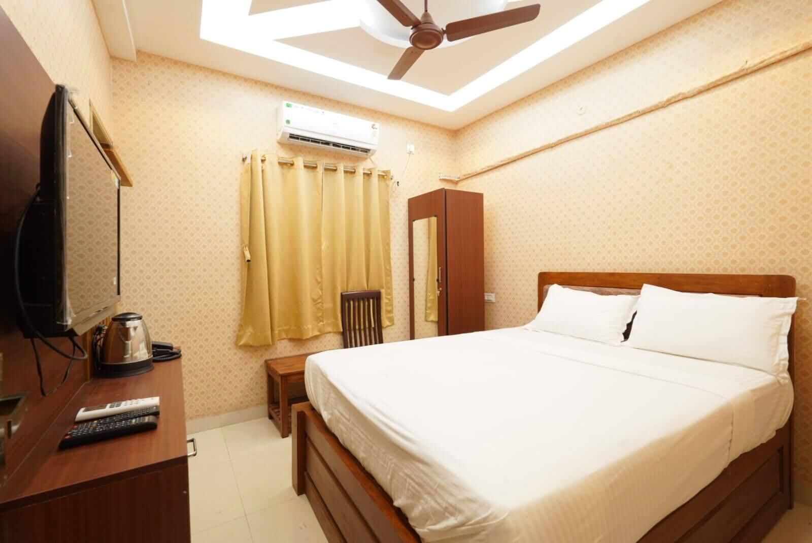 A hotel room with a bed, a desk with a television and remote controls, a chair, a wardrobe, and an air conditioning unit.