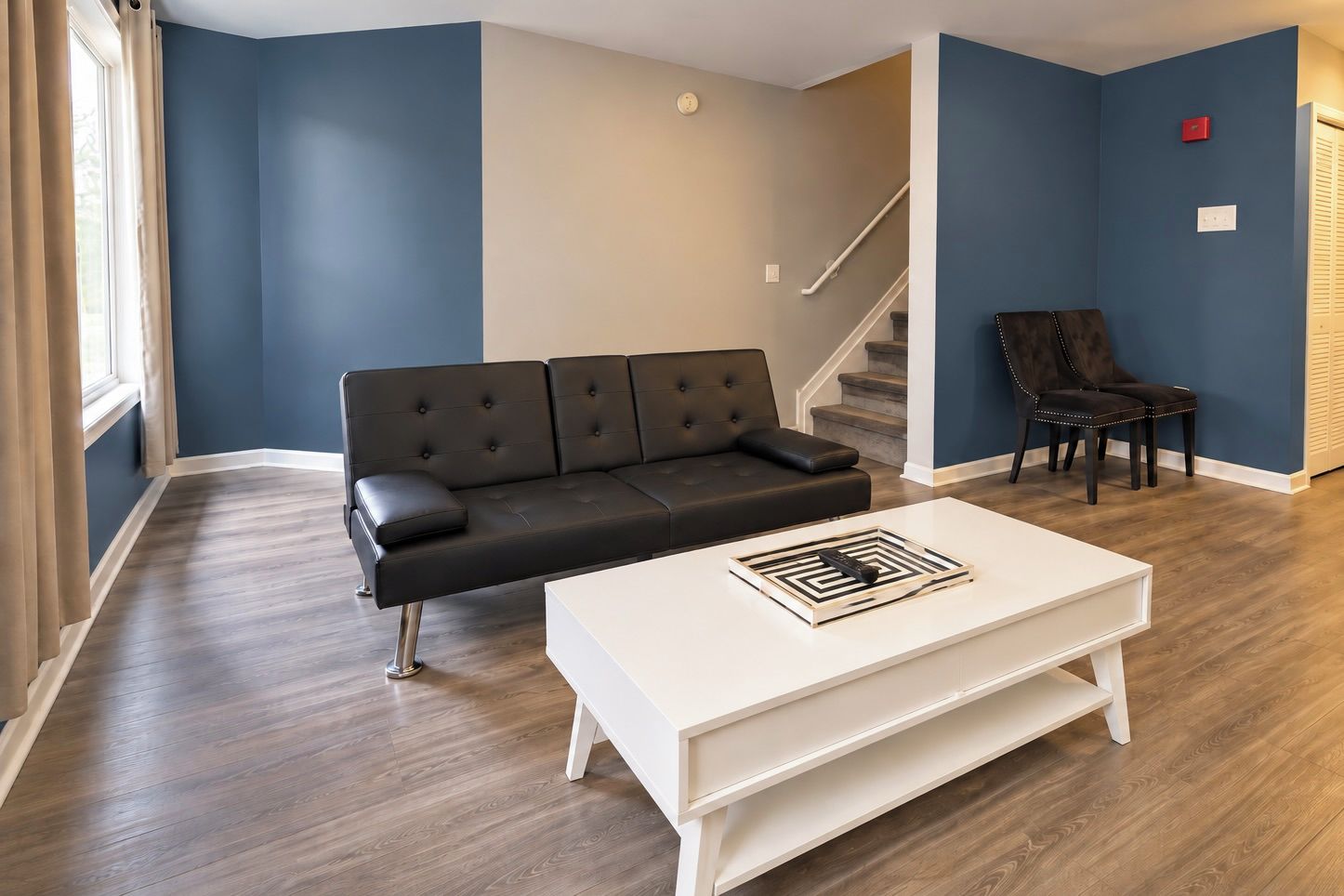 A modern living room with a black leather sofa, a white coffee table, and a staircase in the background.