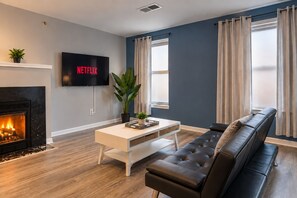 A modern living room with a fireplace, a television mounted on the wall, a black leather sofa, a white coffee table, and a large window with curtains.