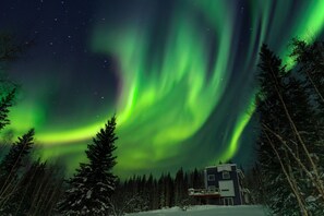 Ein Haus mit einer Terrasse, umgeben von schneebedeckten Bäumen und dem Polarlicht.