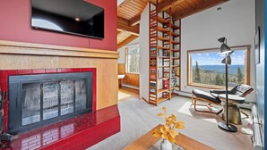 Ein modernes Wohnzimmer mit Kamin, Wandfernseher, Bücherregal, Sessel und Blick ins Freie.