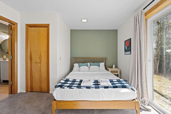 A bedroom with a wooden bed, a checkered blanket, a nightstand, and a window with a view of trees.