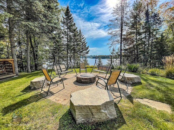 A circular stone fire pit with four chairs, surrounded by a grassy area with trees and a lake in the background. - Sunrise Pines Escape, Butternut Lake w/Hot Tub (Butternut)