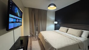 A hotel room with a bed, a TV mounted on the wall, a desk with a lamp, and a window with curtains. - Vive Luxe Condo , Pool & 24/7 Security near Airport (D'Abadie)